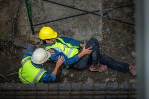 a construction worker tending to another workers who is lying on the ground clutching their leg