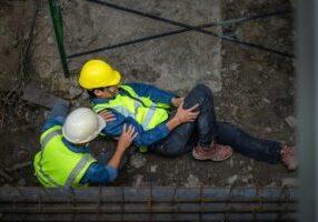 a construction worker tending to another workers who is lying on the ground clutching their leg